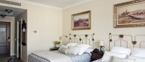 A hotel room with two beds, a wooden door, a nightstand with a lamp, and two framed paintings on the wall.