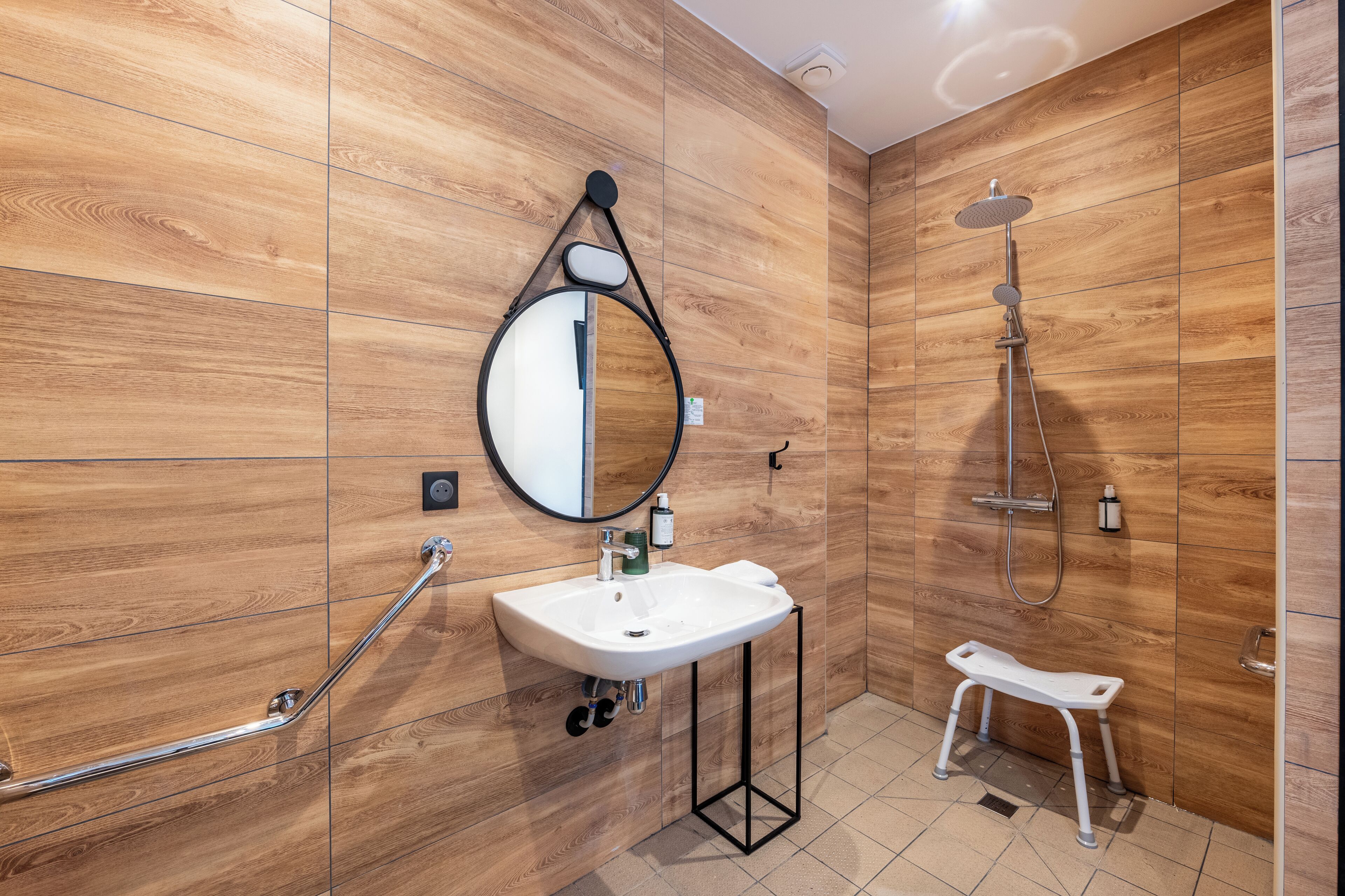 A modern bathroom with a wooden wall, a white sink, a round mirror, a handheld showerhead, and a white step stool.