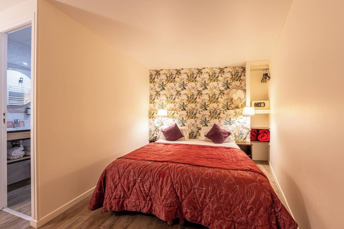 A bedroom with a floral wallpaper headboard, a red bedspread, and a nightstand with a lamp and radio.
