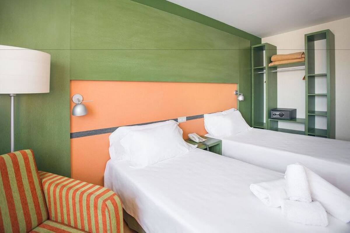A hotel room with two beds, a striped chair, a lamp, and a green shelf with a towel.
