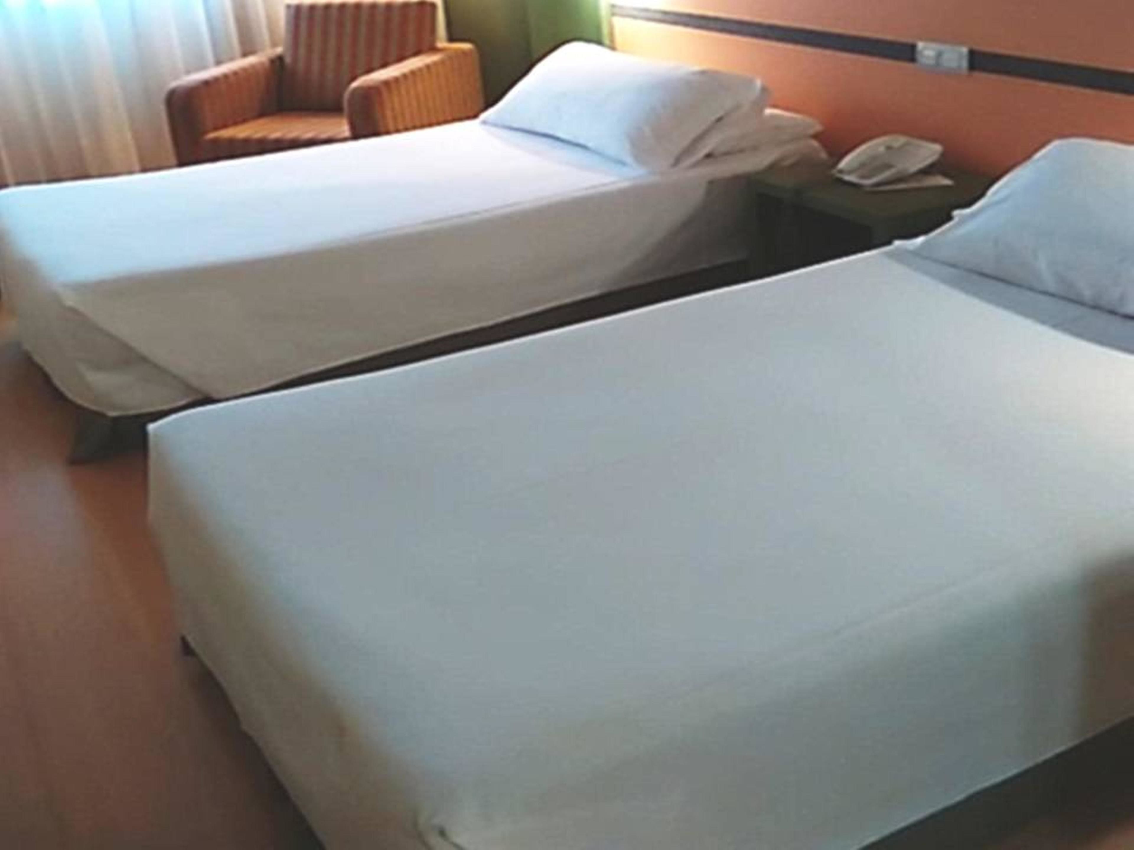 Une chambre d’hôtel avec deux lits simples, une chaise, une table de chevet avec un téléphone et une fenêtre avec des rideaux.