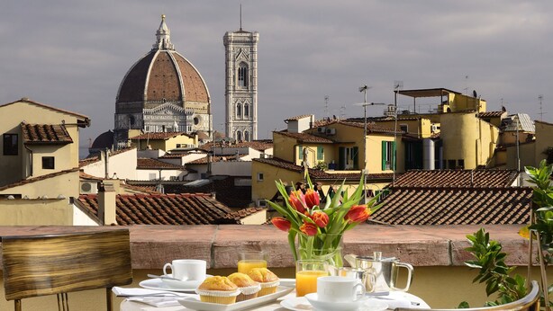 A table with breakfast items and a vase of flowers on a terrace overlooking a cityscape with historic buildings and a tower. - Hotel L'Orologio Firenze (Florence)