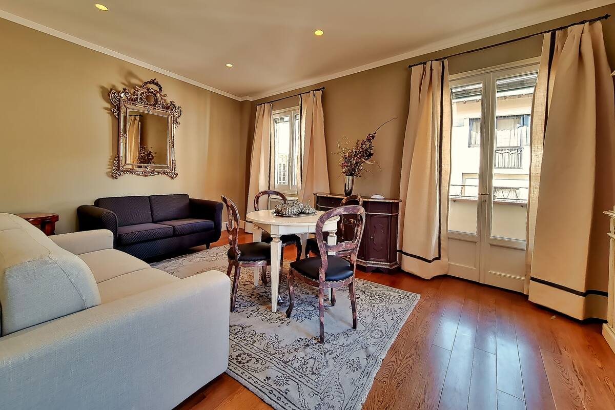 A living room with a sofa, a dining table with chairs, a mirror, and a window with curtains.
