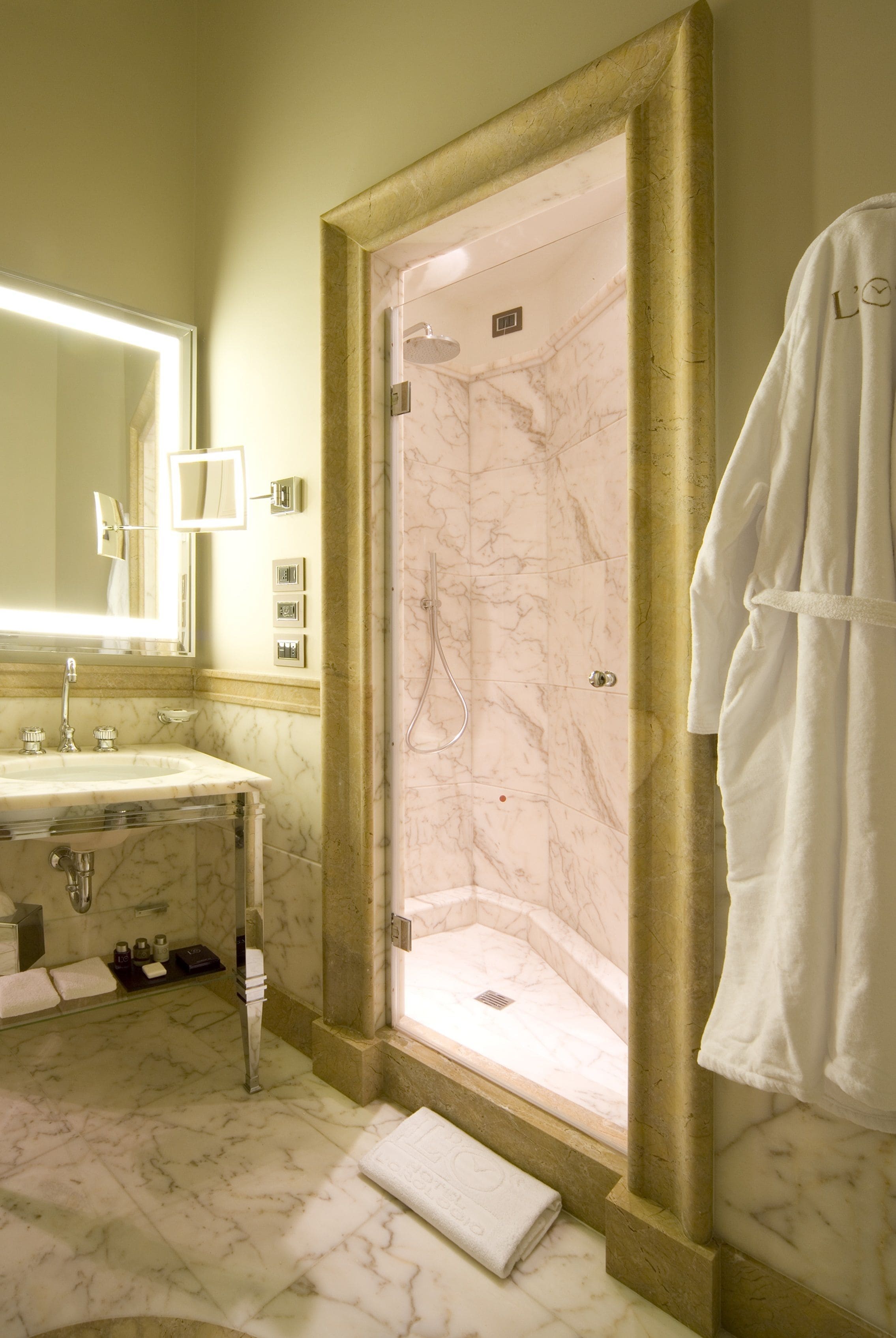 A bathroom with a marble sink, a large mirror, a glass-enclosed shower, and a white robe hanging on the wall.