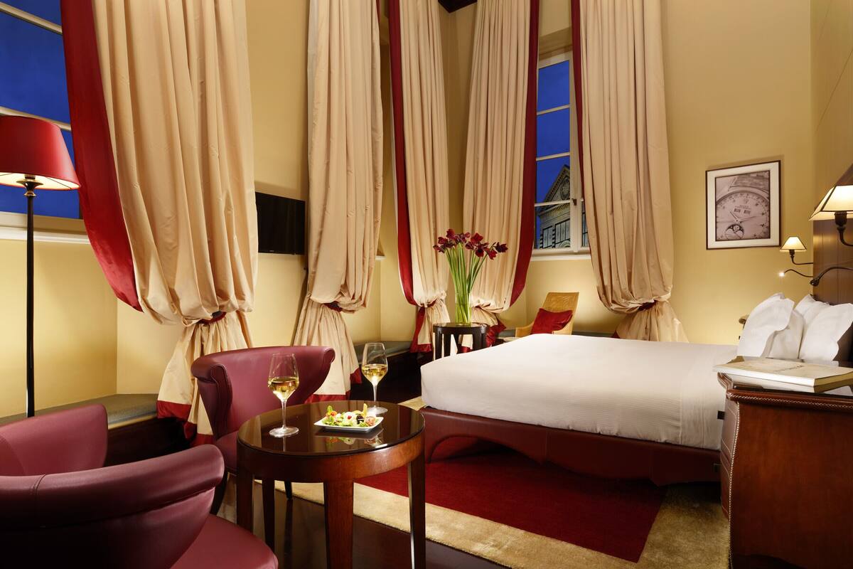 A hotel room with a bed, a desk, a chair, a table with a wine glass, and curtains.