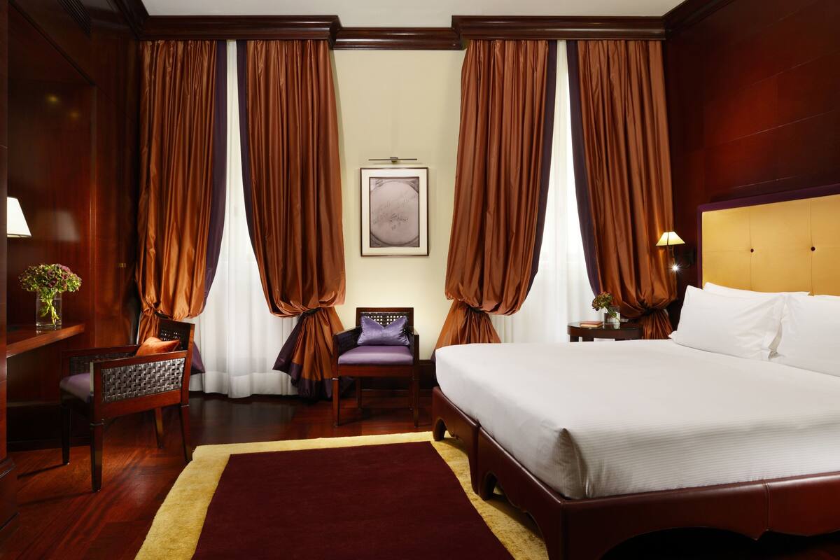 A hotel room with a large bed, wooden headboard, a nightstand with a lamp, a chair, and a framed picture on the wall.