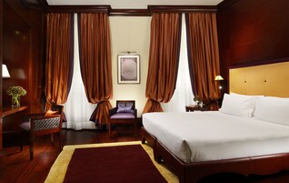 Una camera d'albergo con un letto grande, una testiera in legno, un comodino con una lampada, una sedia e un quadro incorniciato appeso al muro.