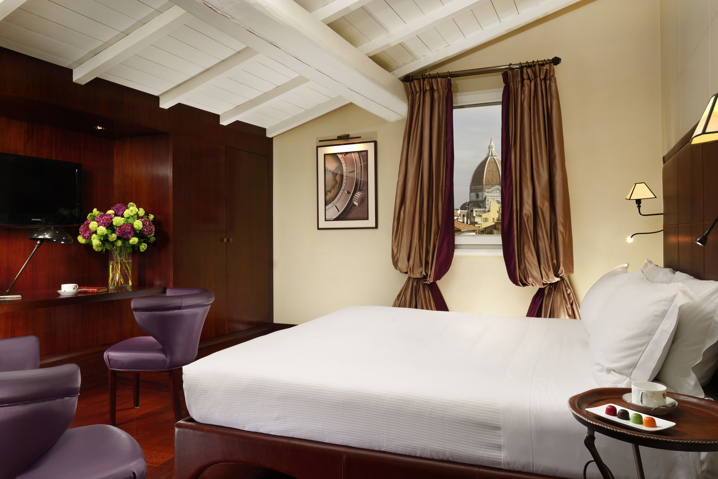A hotel room with a large bed, a desk with a flat-screen TV, purple chairs, a vase with flowers, and a window with curtains.
