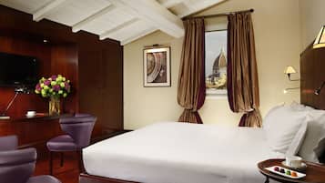 A hotel room with a large bed, a desk with a flat-screen TV, purple chairs, a vase with flowers, and a window with curtains.