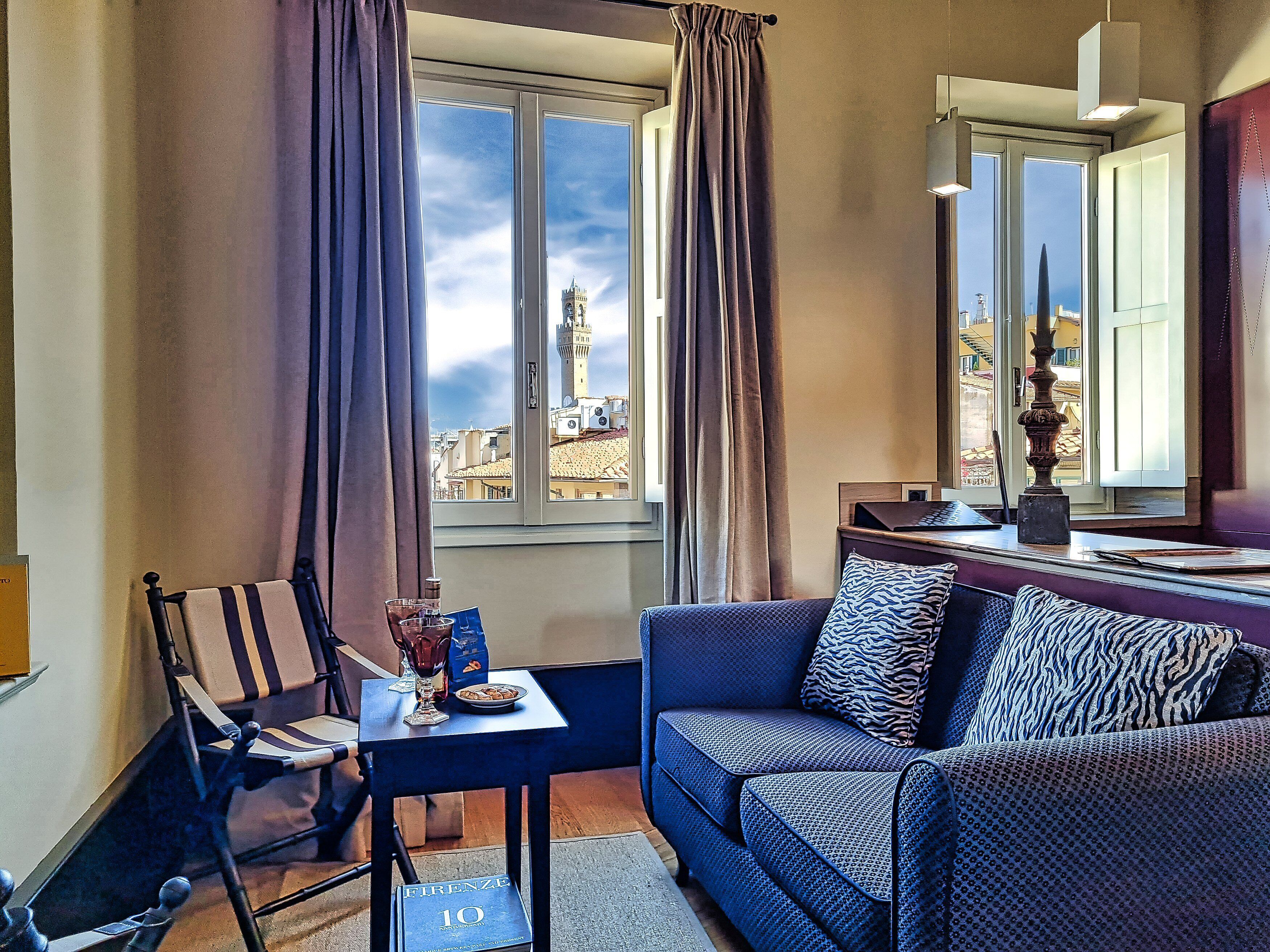 Una habitación con vistas a una torre, un sofá, una silla, una mesa con un vaso y un libro, y una ventana con cortinas.