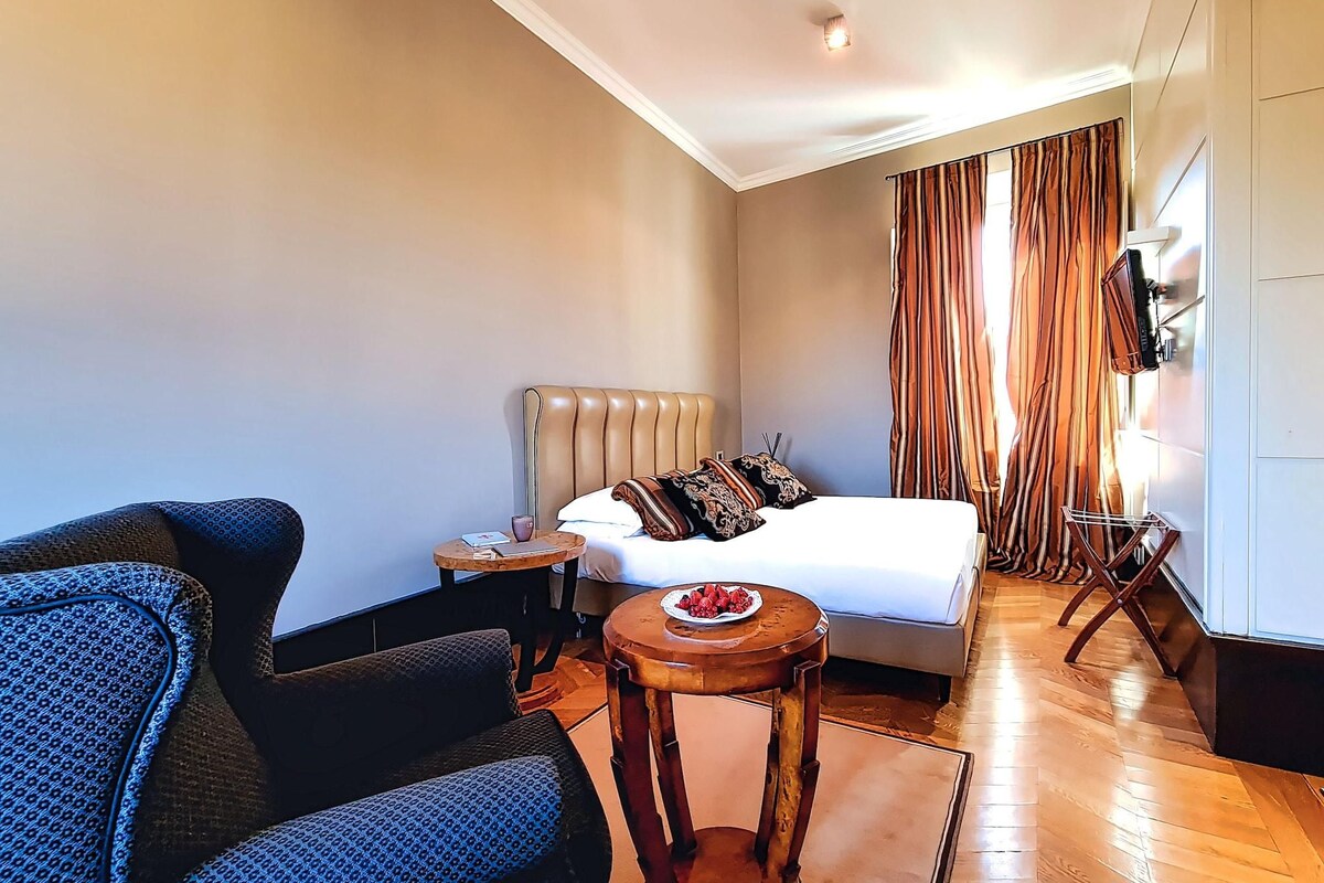 A hotel room with a bed, a small round table with fruit, a sofa, and a TV on the wall.