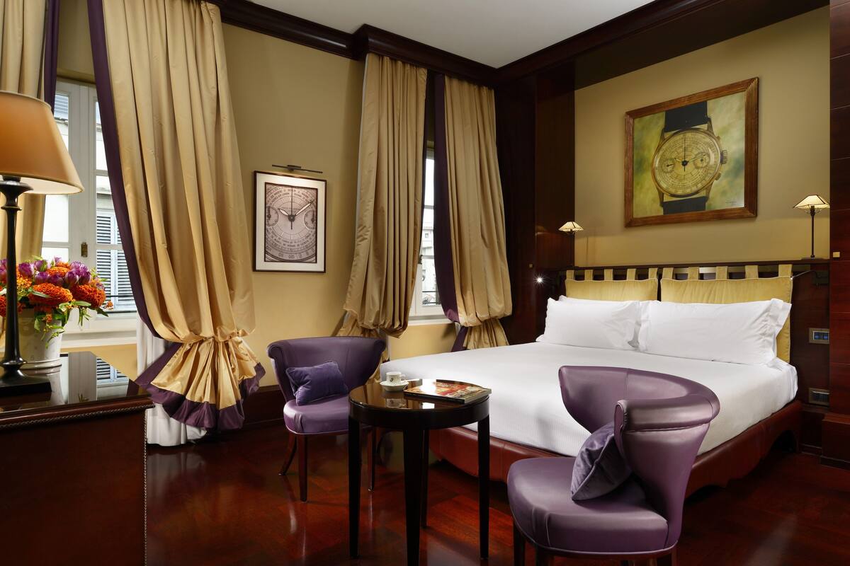 A hotel room with a large bed, two purple armchairs, a small table with a book and a cup, a vase with flowers, and a painting on the wall.