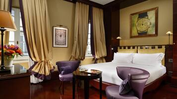 Una camera d'albergo con un letto grande, due poltrone viola, un tavolino con un libro e una tazza, un vaso con dei fiori e un quadro appeso al muro.