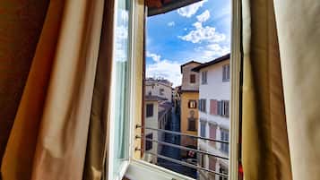 Una ventana con vistas a una calle estrecha con edificios, una barandilla metálica y cielo despejado.