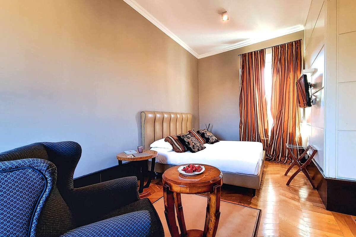 A hotel room with a bed, a small round table with fruit, a sofa, and a TV on the wall.
