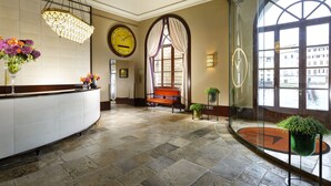 A hotel lobby with a reception desk, a clock, a bench, and a large window offering a view of the city. - Hotel L'Orologio Firenze (Florence)