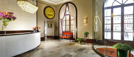 A hotel lobby with a reception desk, a clock, a bench, and a large window offering a view of the city.