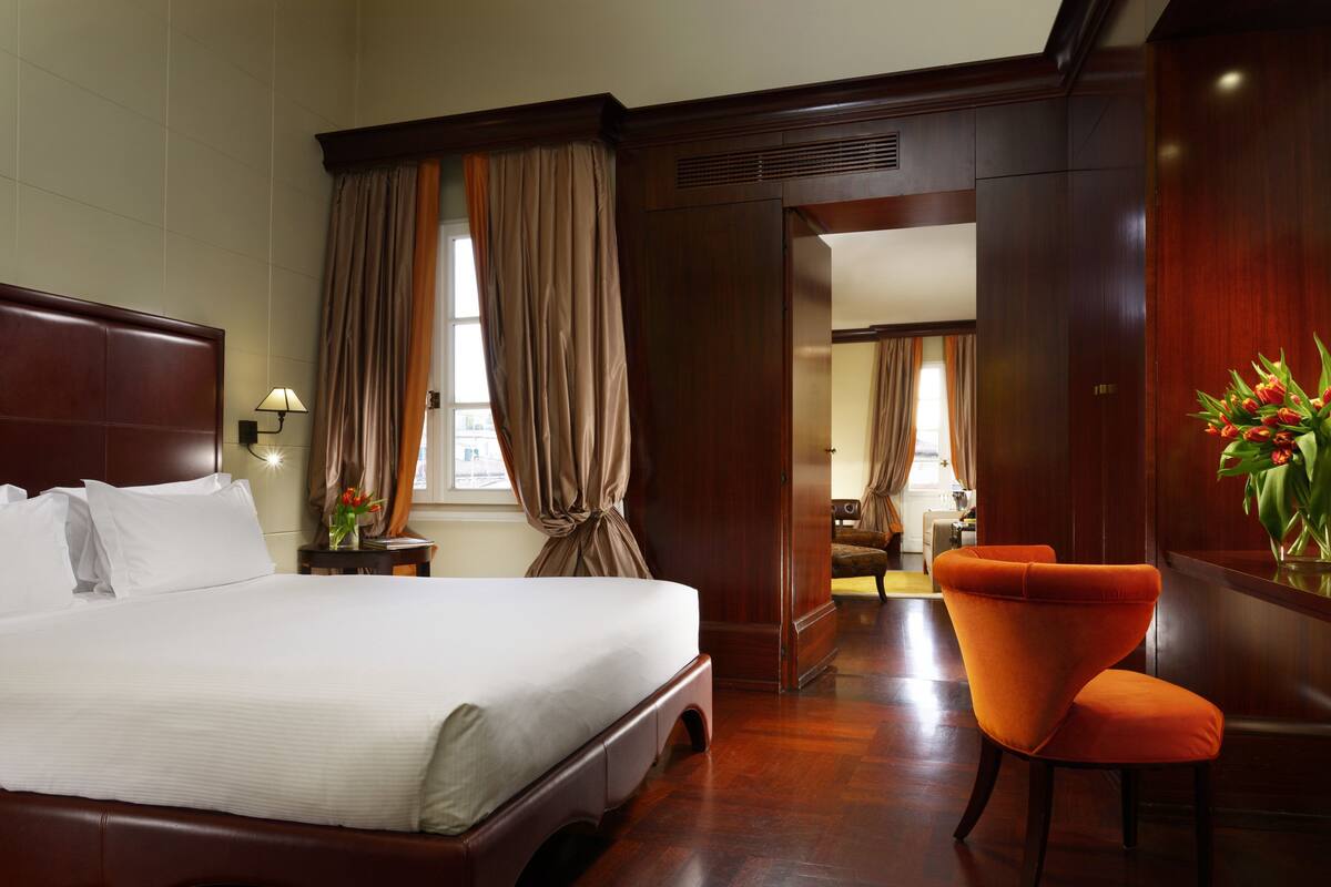 A hotel room with a large bed, an orange armchair, a wooden wardrobe, and a mirror reflecting the room.