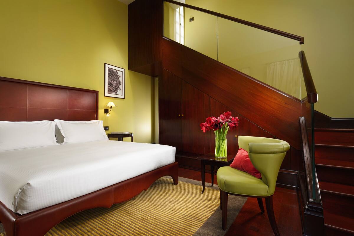 A hotel room with a large bed, a wooden staircase, a small table with a vase of flowers, and a green chair.