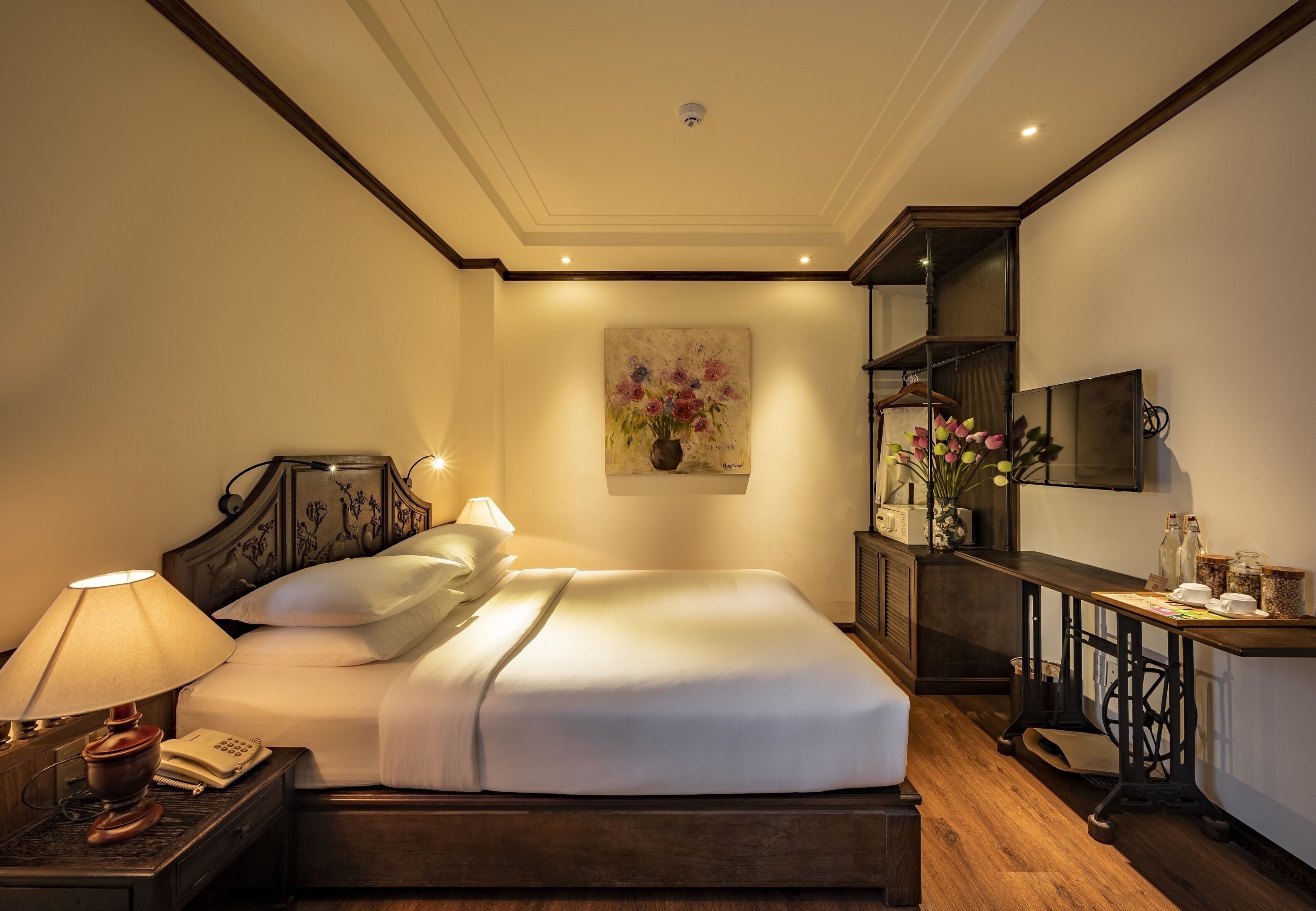 A well-lit bedroom with a large bed, bedside table with a lamp, a painting on the wall, and a TV on a stand.