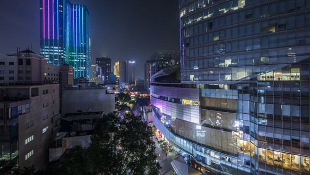 Une vue nocturne d’une ville, avec des immeubles modernes de grande hauteur, dont un est illuminé par des néons verticaux.