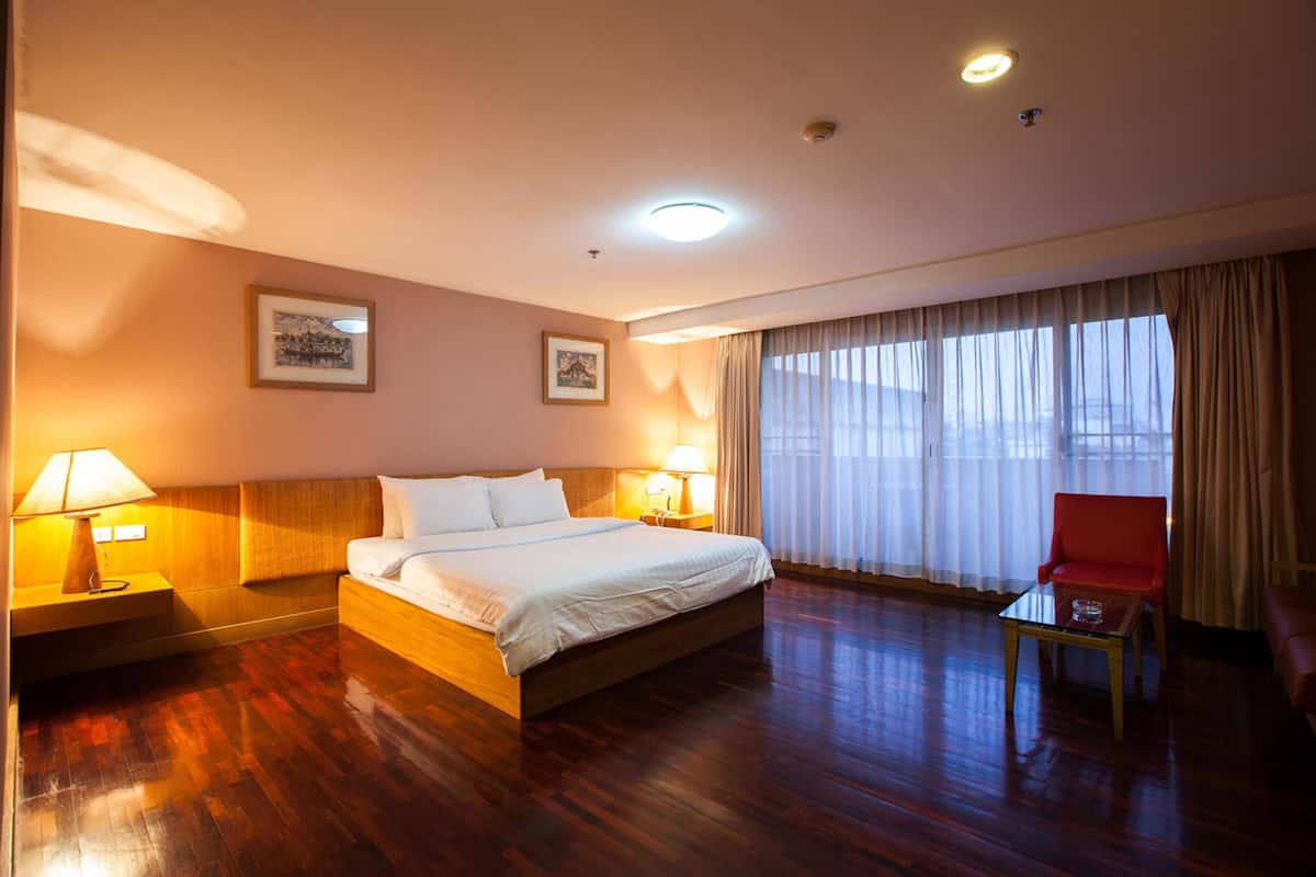 Habitación de hotel con una cama de madera, dos lámparas de noche, un sillón rojo y un ventanal con cortinas traslúcidas.