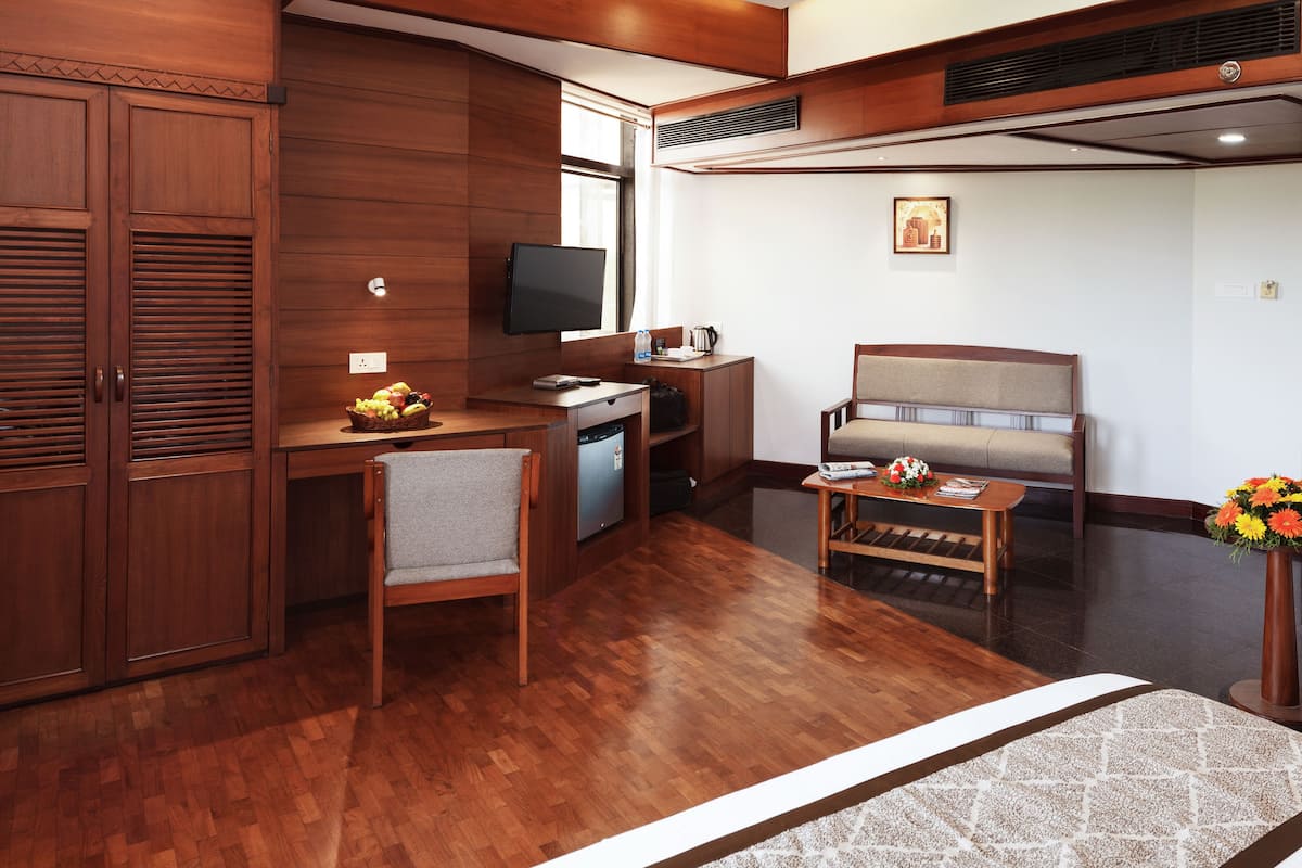 A compact hotel room with a wooden desk, a television, a sofa, and a small table with a vase of flowers.