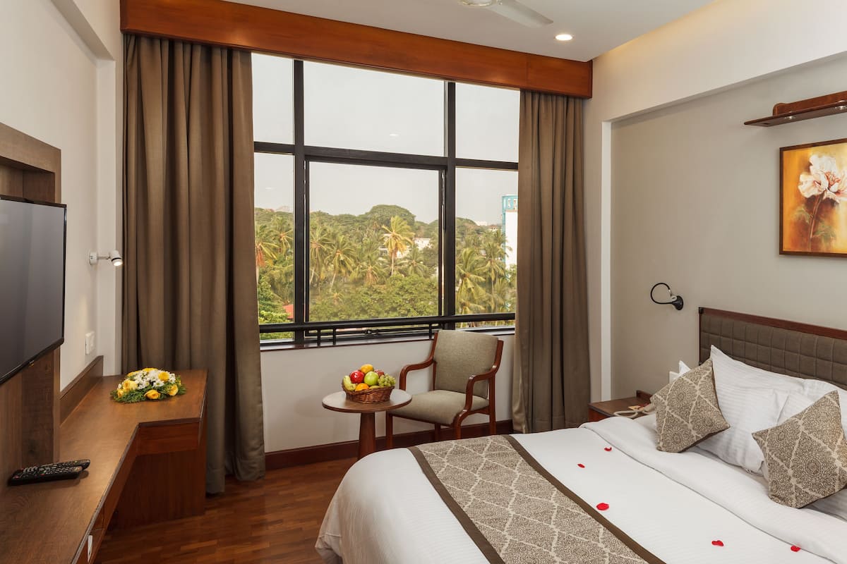 A hotel room with a large window, a bed with patterned pillows, a small table with fruit, and a TV on a wooden desk.