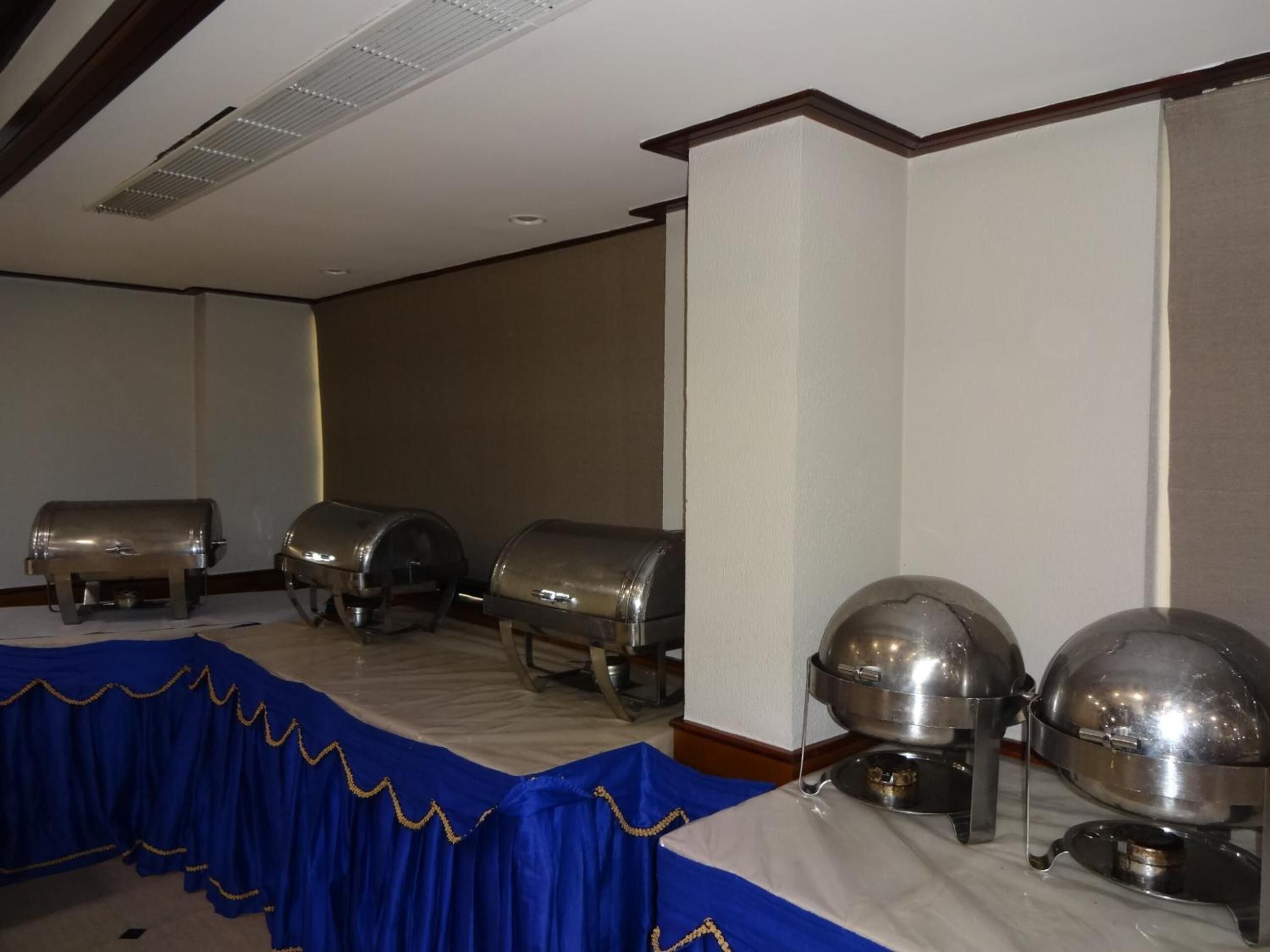 A room with a long table covered by a blue tablecloth, lined with silver buffet trolleys and a large silver urn.
