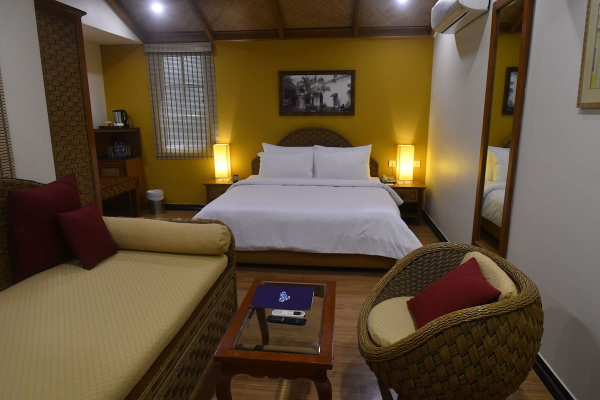 Habitación de hotel con cama, dos sofás, una mesita con un control remoto y una silla de mimbre.