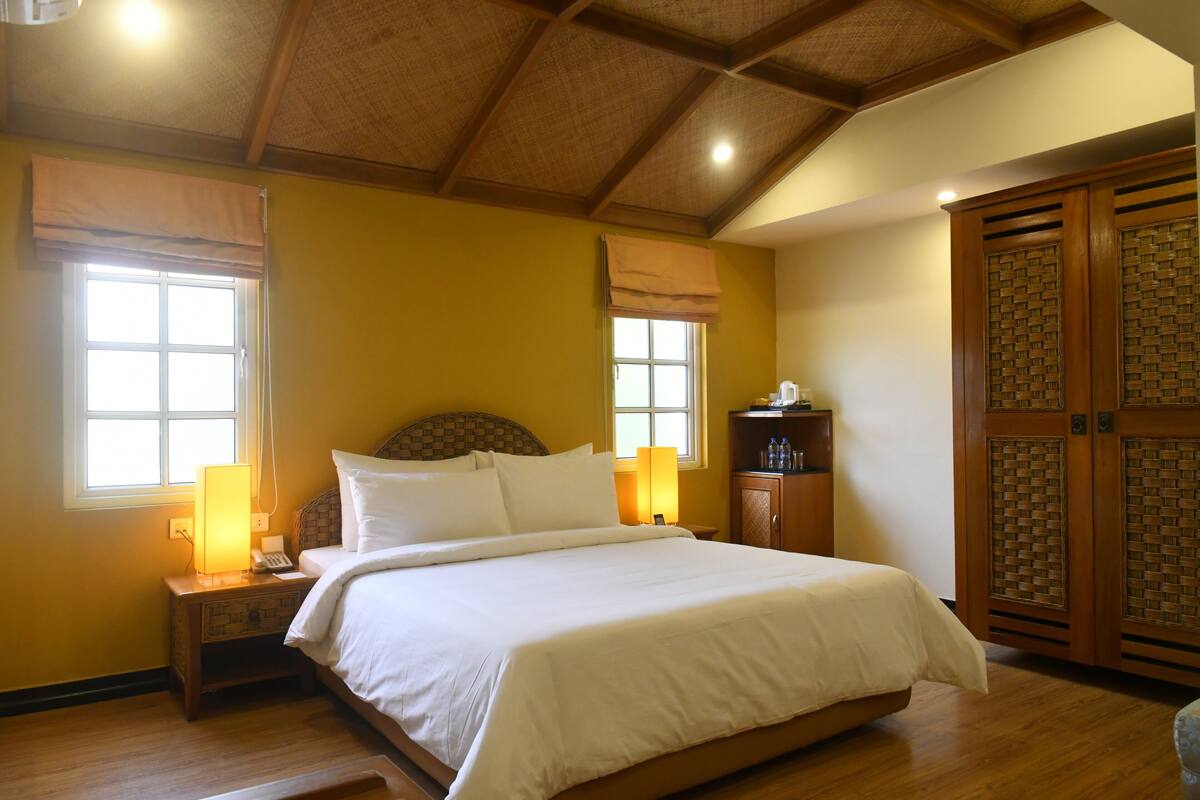Un dormitorio con una cama grande, cabecera de madera, dos lámparas de noche, una mesita de noche, un armario y una ventana con vistas.