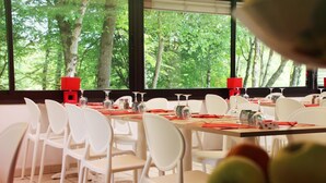 Un espace repas avec des chaises et des tables blanches, des serviettes rouges et une vue sur les arbres à travers de grandes fenêtres.