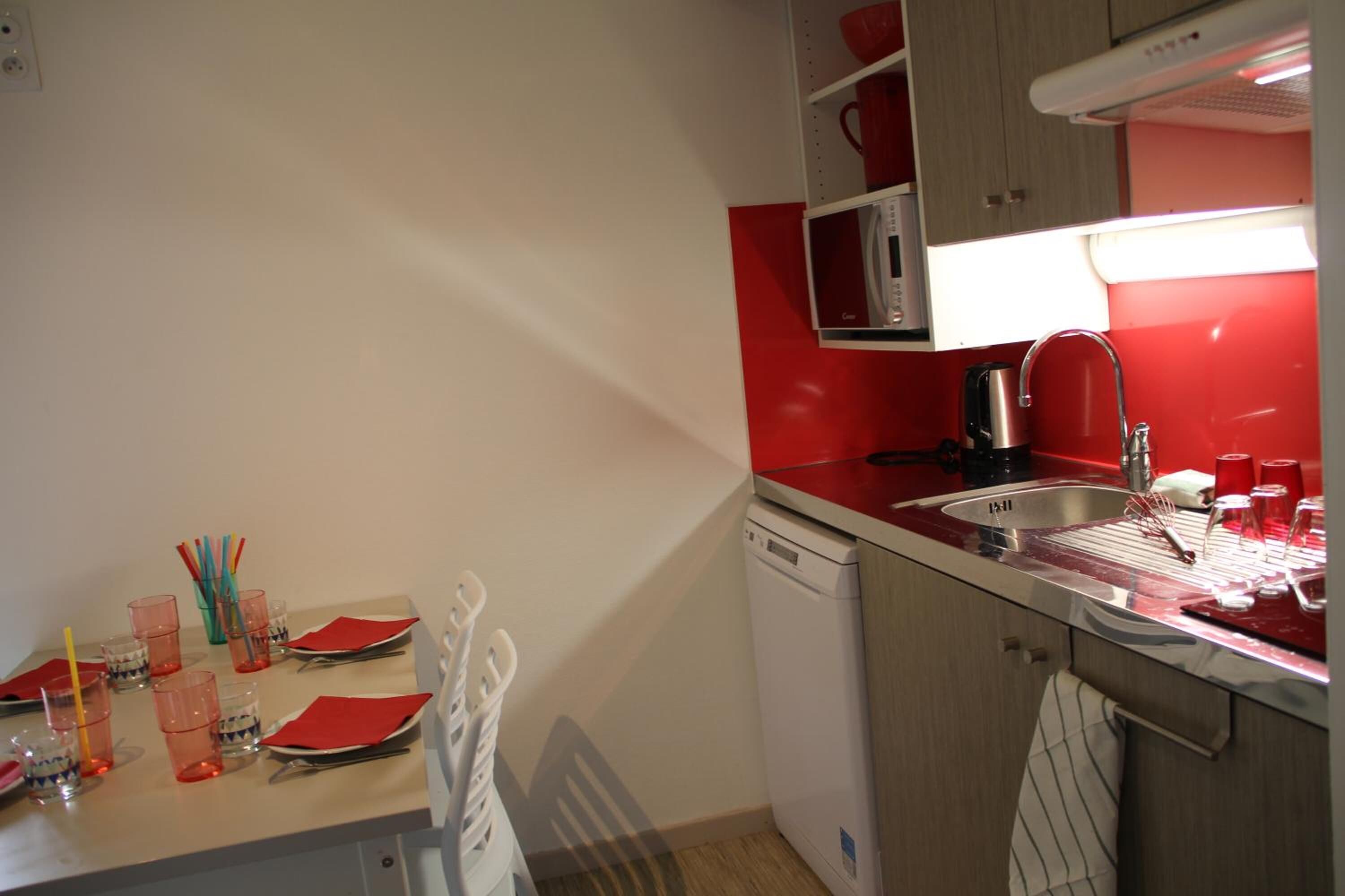 Una cocina compacta con un lavabo, un microondas y un lavavajillas. Hay armarios rojos y blancos, y una mesa dispuesta con servilletas y vasos rojos.