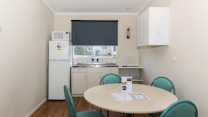 A compact kitchen with a microwave, refrigerator, and sink. There is a dining table with chairs and a window with blinds.