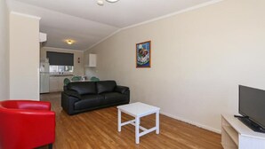 A compact living space with a black leather sofa, a red chair, a white coffee table, and a TV on a white stand. A kitchen area is visible in the background.