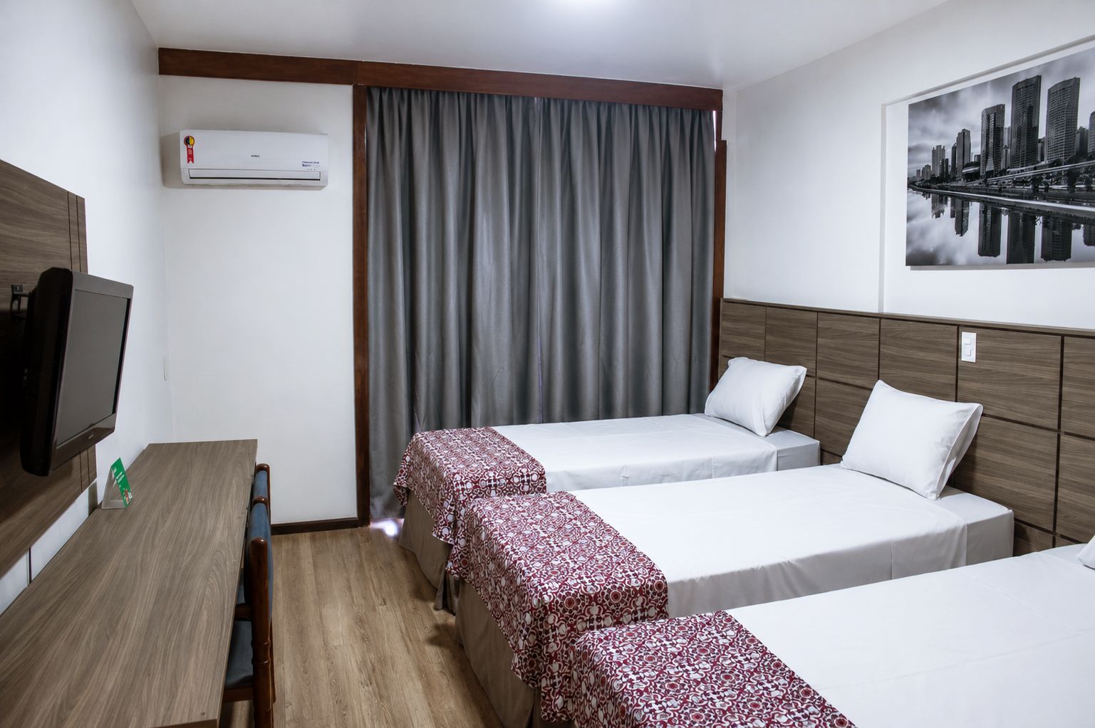 A hotel room with two beds, a desk with a television, an air conditioner, and a cityscape view.