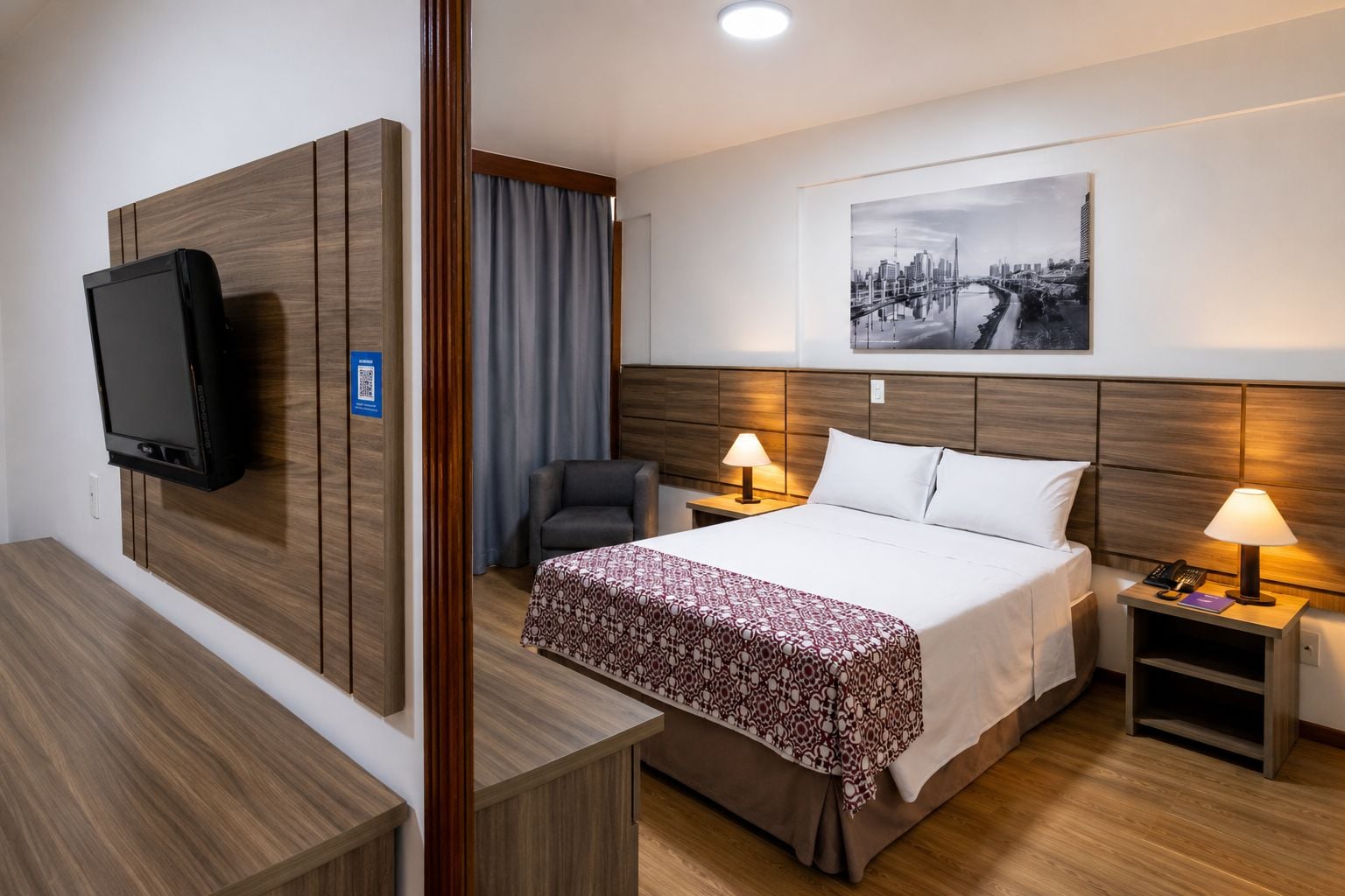 A hotel room with a bed, bedside tables, a TV, a chair, and a cityscape painting on the wall.