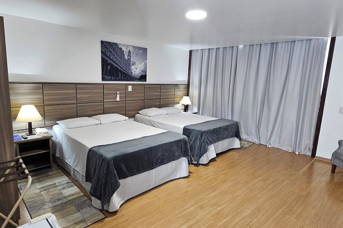 A hotel room with two beds, a wooden headboard, a painting, a desk with a lamp, and a grey sofa.