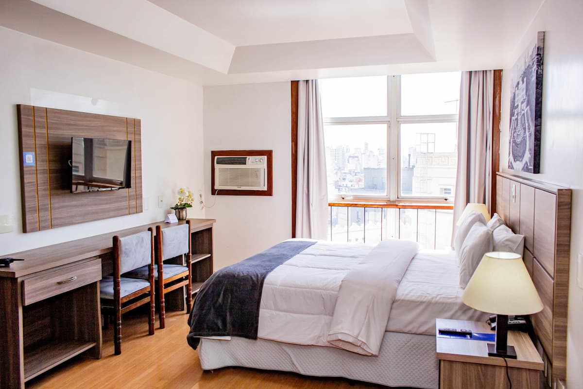 A hotel room with a bed, a desk, a chair, a TV, an air conditioner, and a window with a city view.