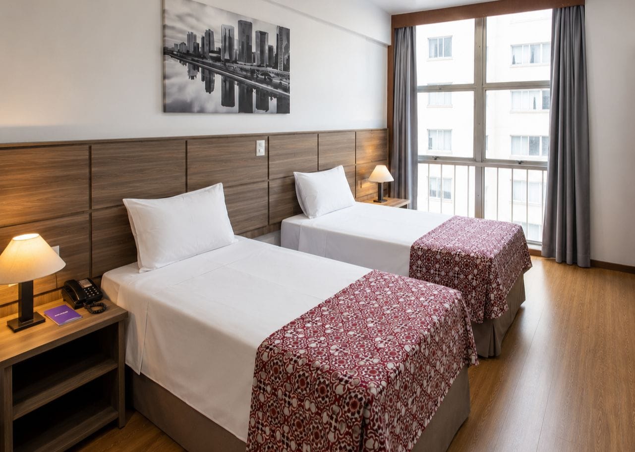 Une chambre d’hôtel avec deux lits, une œuvre d’art représentant un paysage urbain accrochée au mur, une fenêtre avec des rideaux et une table de chevet avec une lampe et un téléphone.