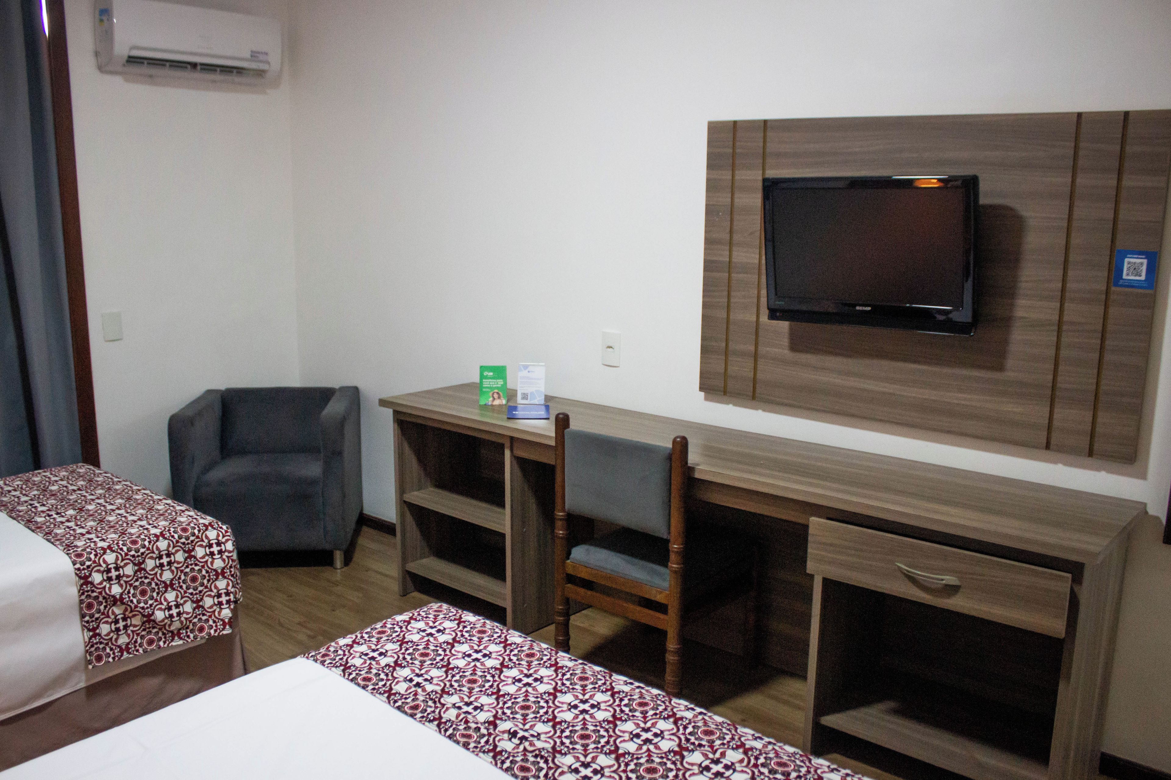 A hotel room with a bed, a desk with a chair, a television mounted on the wall, and a small shelf.