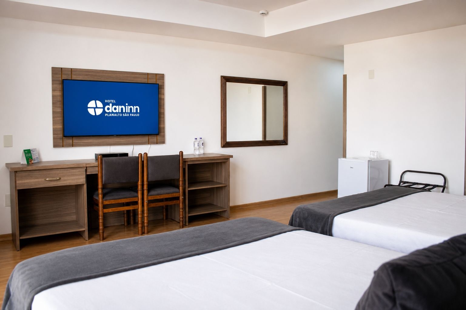 A hotel room with two beds, a desk with a chair, a television, a mirror, and a refrigerator.
