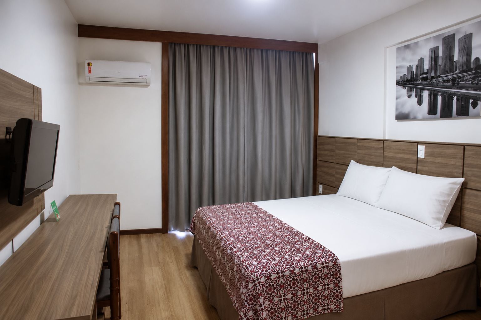 A hotel room with a bed, a desk with a television, an air conditioner, and a cityscape view.