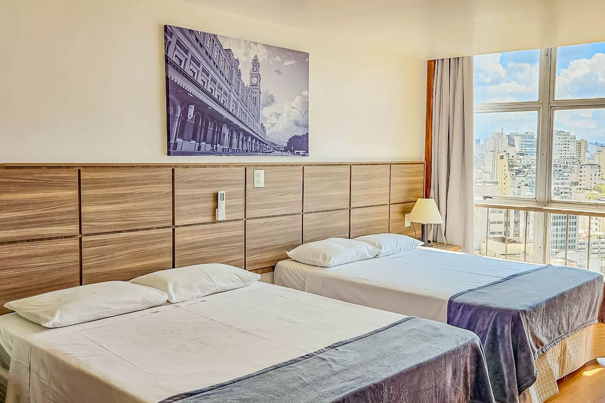 A hotel room with two beds, a large window with a city view, a painting on the wall, and a bedside table with a lamp.