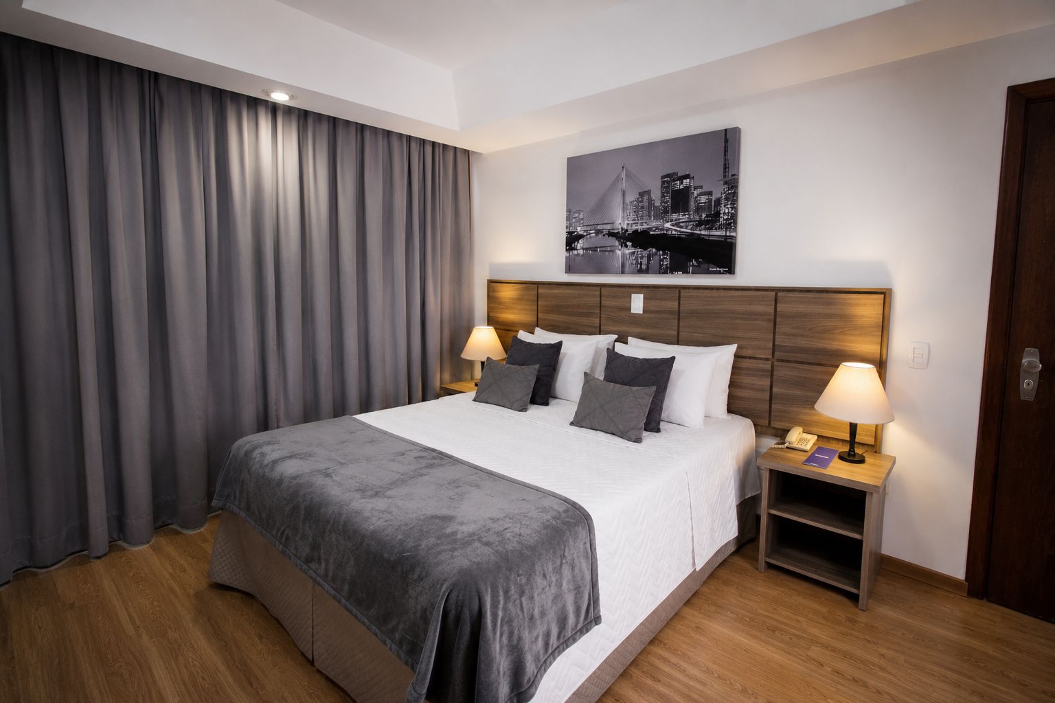 Habitación de hotel con una cama grande, dos mesitas de noche con lámparas, un cuadro de un paisaje urbano en la pared y una puerta de madera.