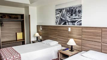 Camera d'albergo con due letti, una testiera in legno, un quadro con una veduta cittadina, un comodino con una lampada e un armadio.