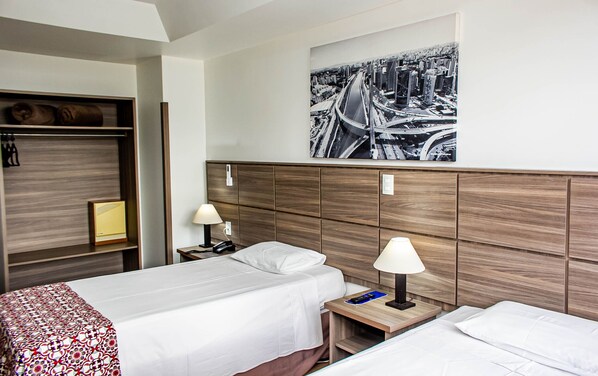 A hotel room with two beds, a wooden headboard, a cityscape painting, a bedside table with a lamp, and a wardrobe.