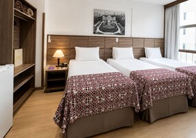 Quarto de hotel com duas camas, mesa de cabeceira, abajur, quadro com vista urbana e janela com cortinas.