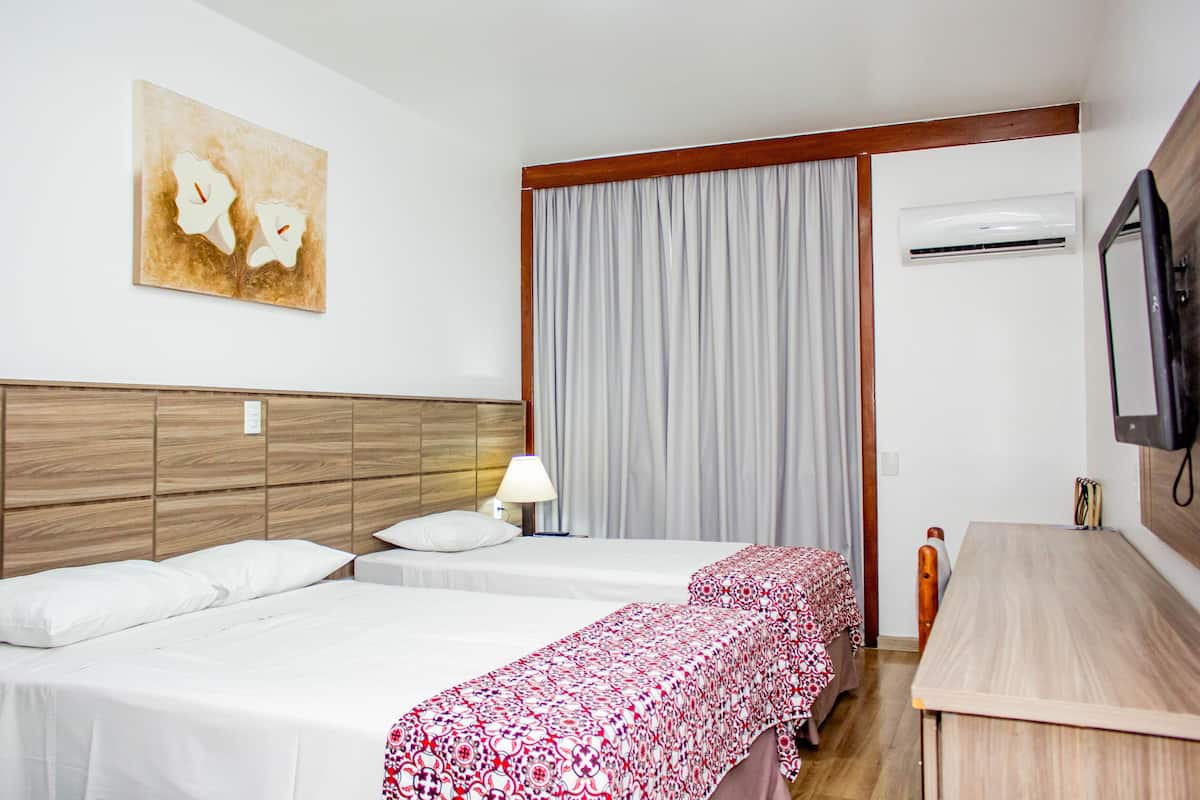 A hotel room with two beds, a wooden headboard, a large window with curtains, an air conditioning unit, a bedside table, and a television.