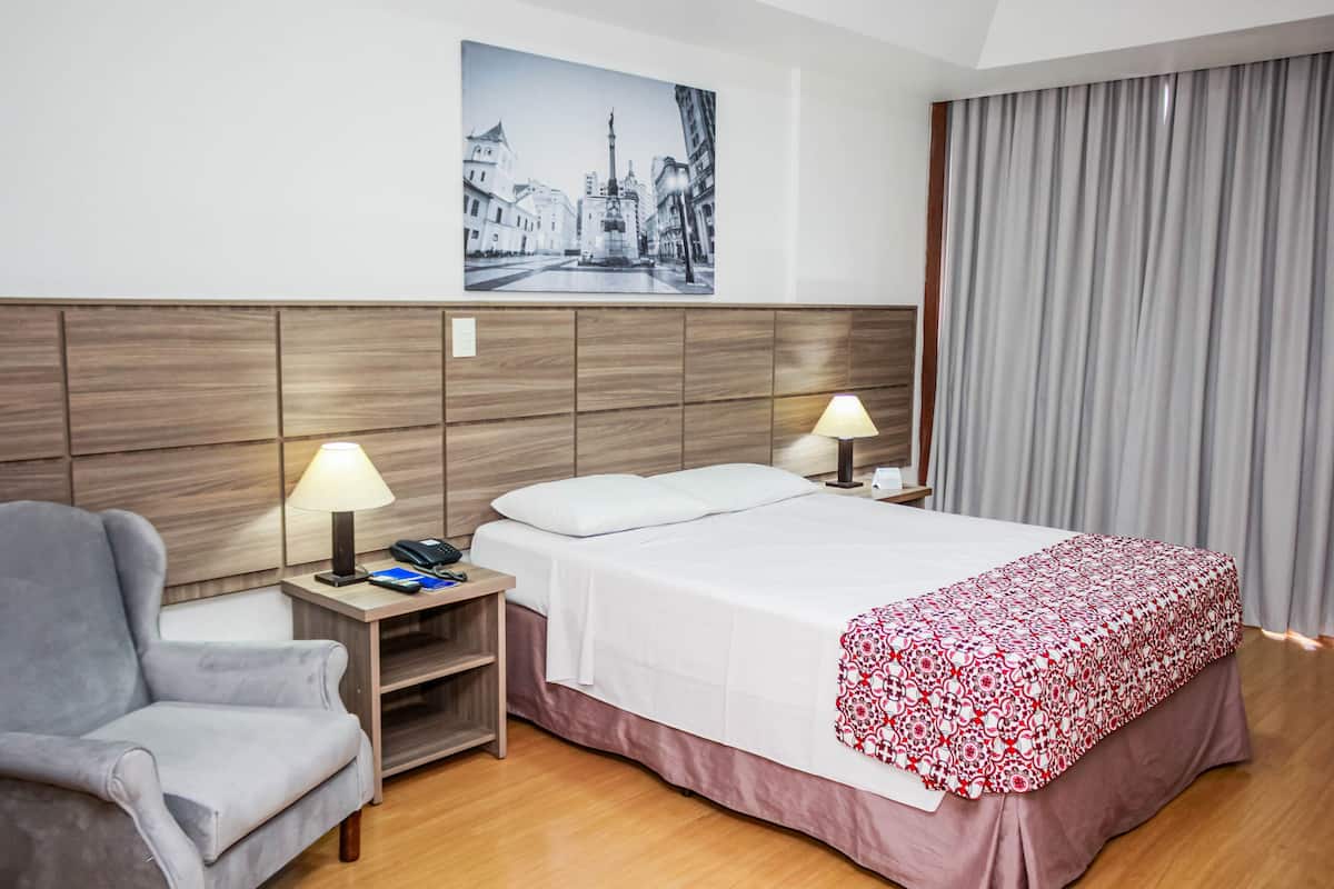 A hotel room with a bed, two bedside lamps, a chair, a nightstand, a framed picture on the wall, and a window with curtains.